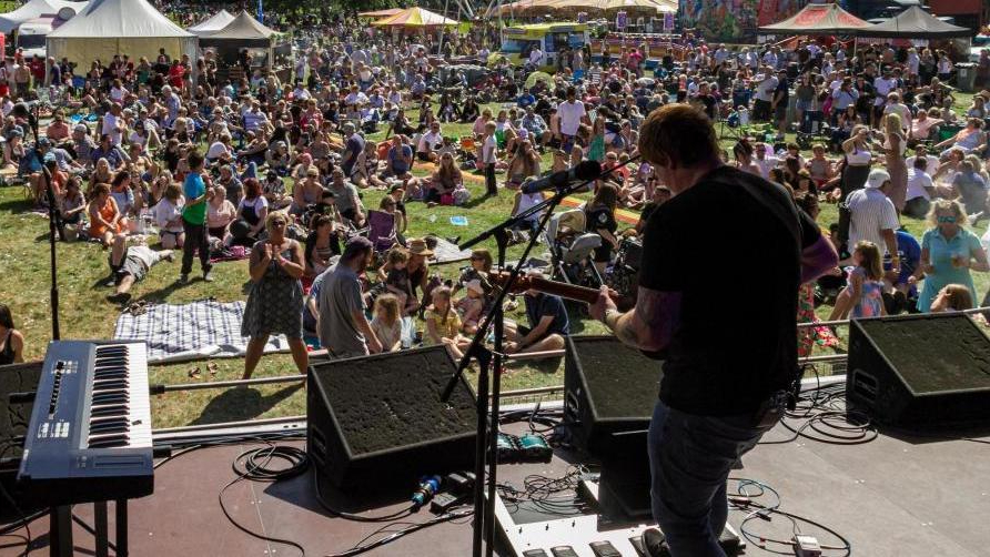 An outdoor festival scene with a crowd of people gathered in front of a stage.