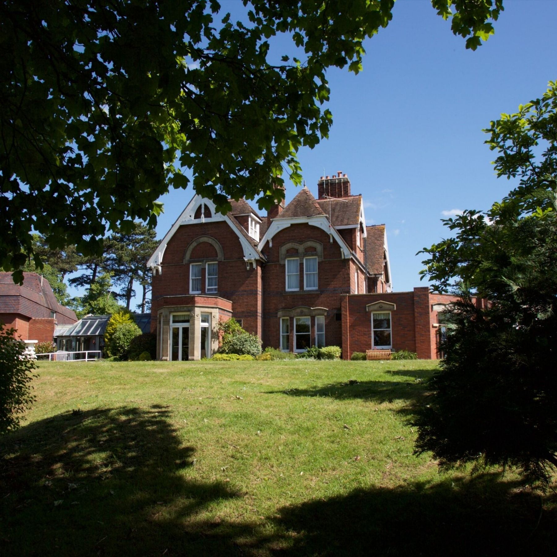 An exterior view of the red-brick Victorian manor house framed by green trees and a lawn.