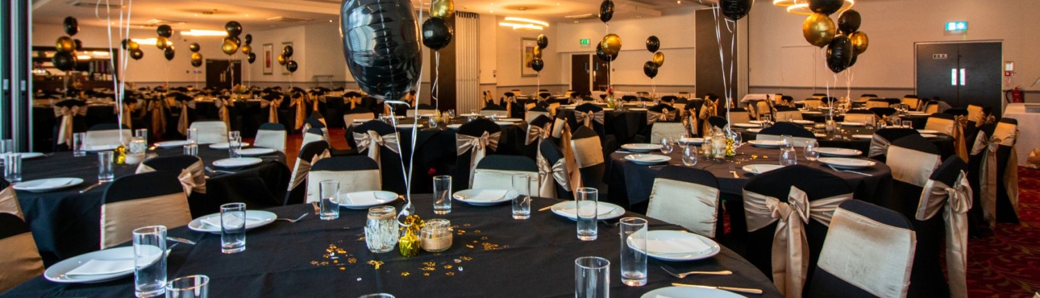A banquet hall set with round tables, black linens, and gold balloon centerpieces.