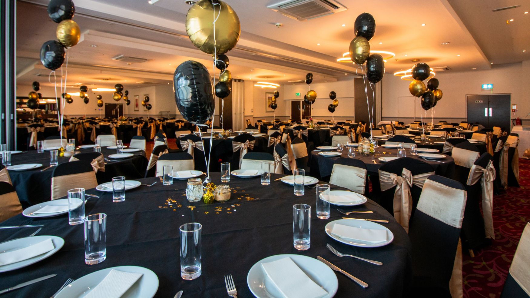 A banquet hall set with round tables, black linens, and gold balloon centerpieces.