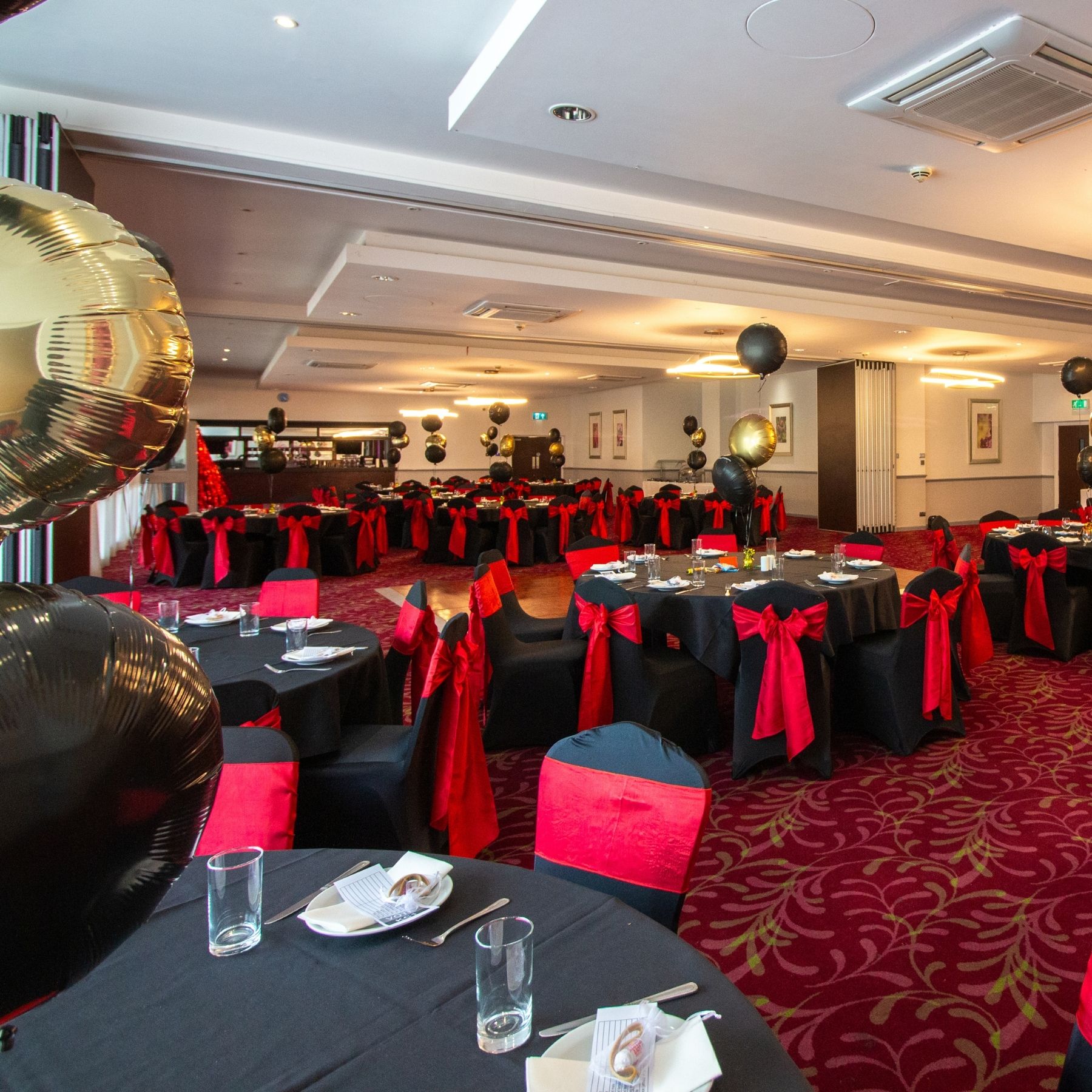 A large banquet hall set with round tables, red chair sashes, and black tablecloths.