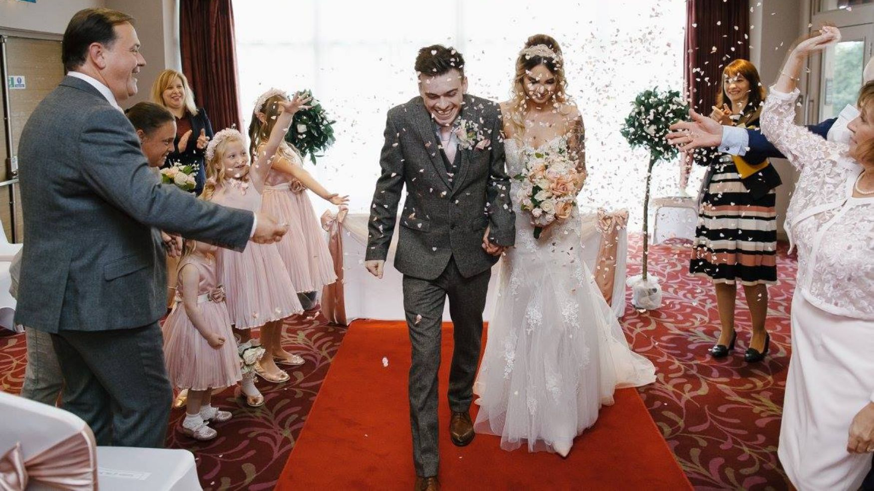 A bride and groom walking down a red carpet aisle after their ceremony, being cheered by guests.