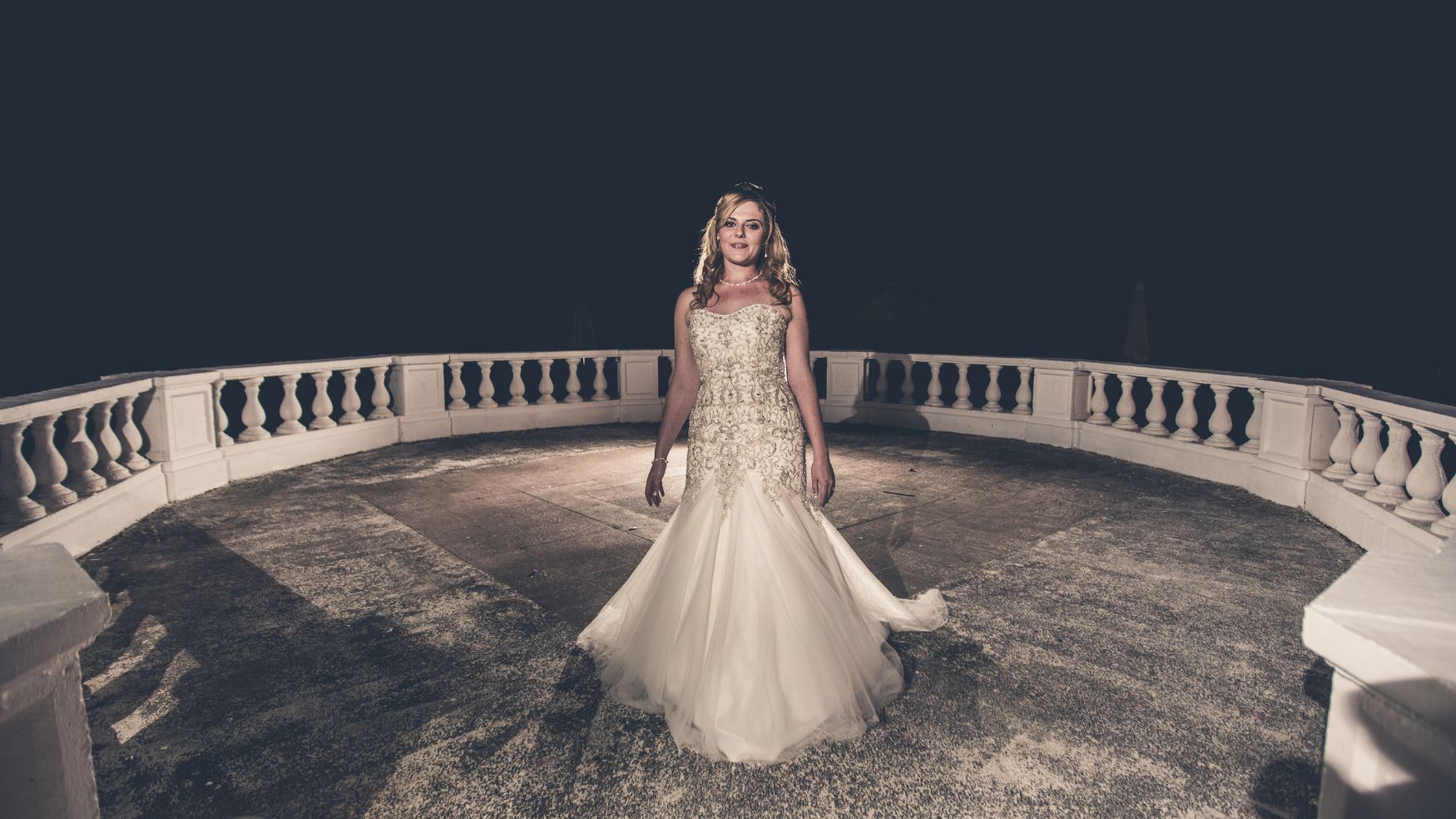 A bride in a flowing gown standing alone on a balcony or terrace at night.