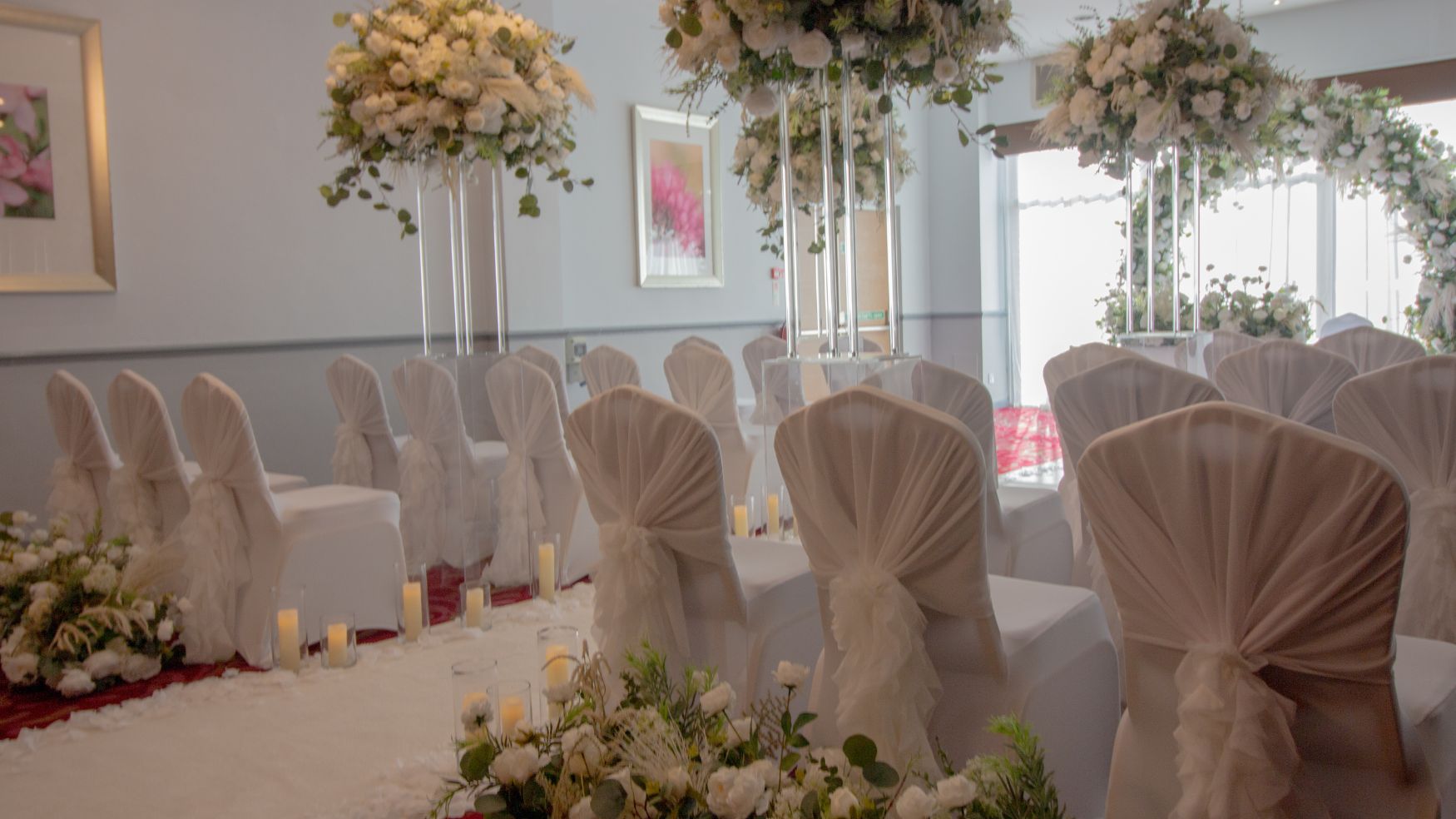 A ceremony room with white chair covers and floral arrangements at the end of the aisles.