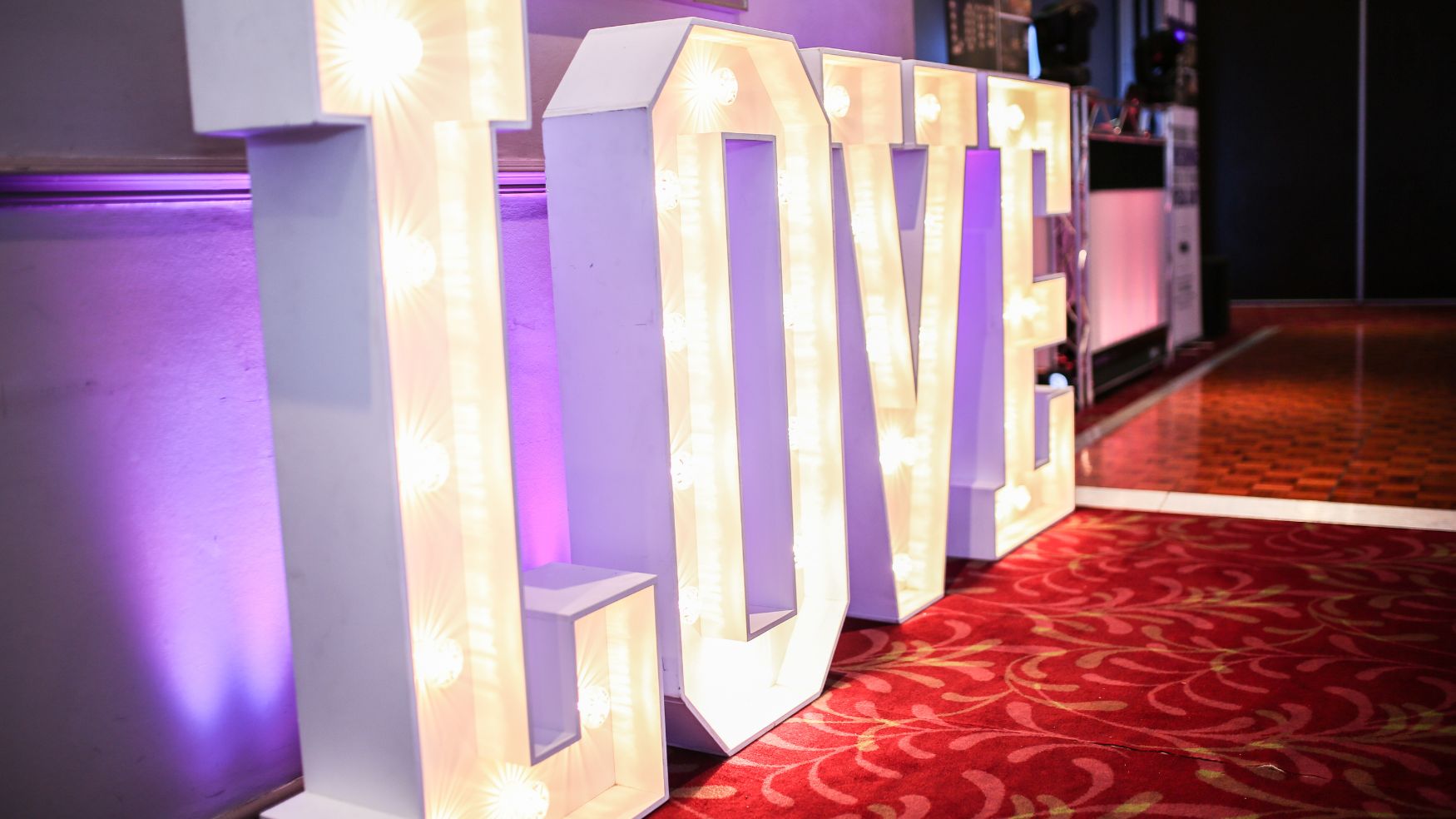 A set of tall, white illuminated "LOVE" marquee letters standing on a red patterned carpet.