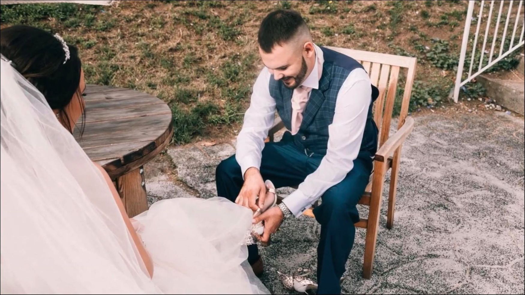 A groom sitting on a wooden chair outdoors, looking down while adjusting the bride's shoes.