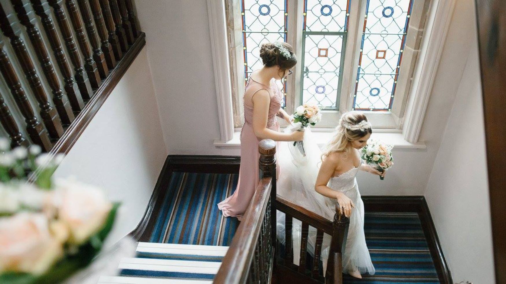 A bride descending a classic wooden staircase near a stained-glass window.