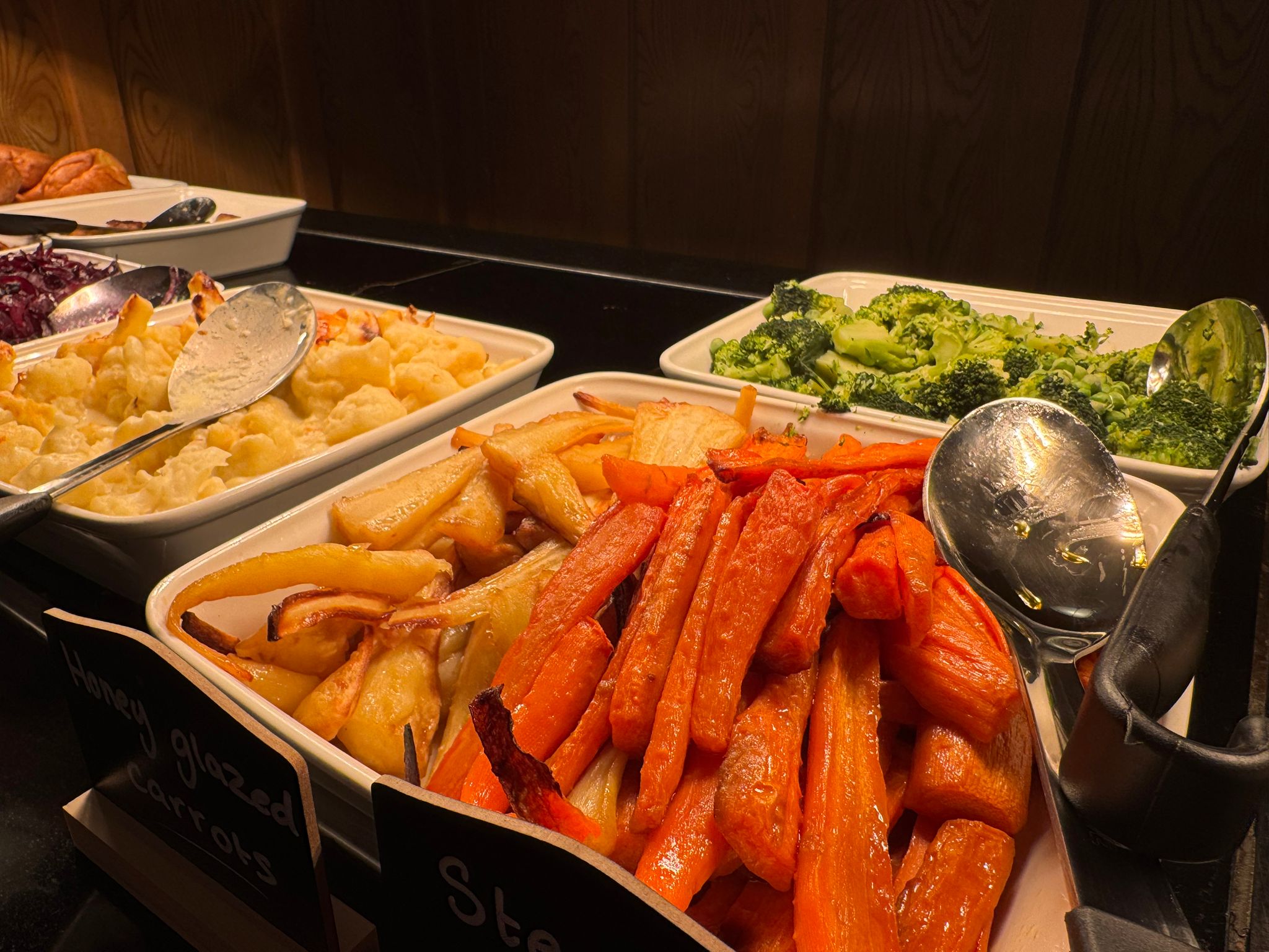 A buffet tray filled with vibrant, honey-glazed roasted carrots and parsnips, with other vegetable trays in the background.