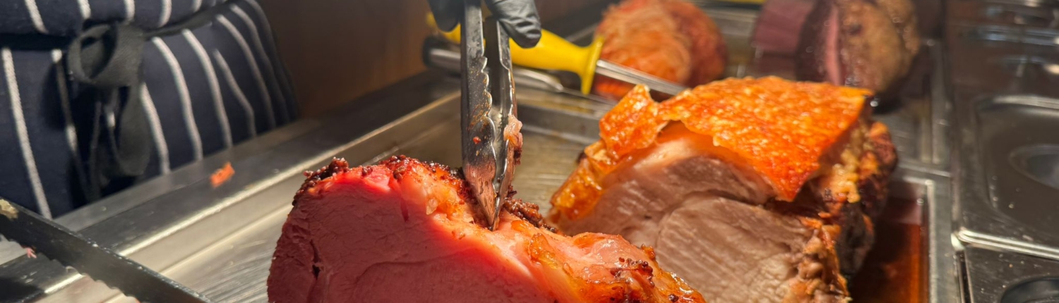 A chef’s hands wearing black gloves, using a carving fork and knife to slice into a succulent, pink roasted joint of meat.