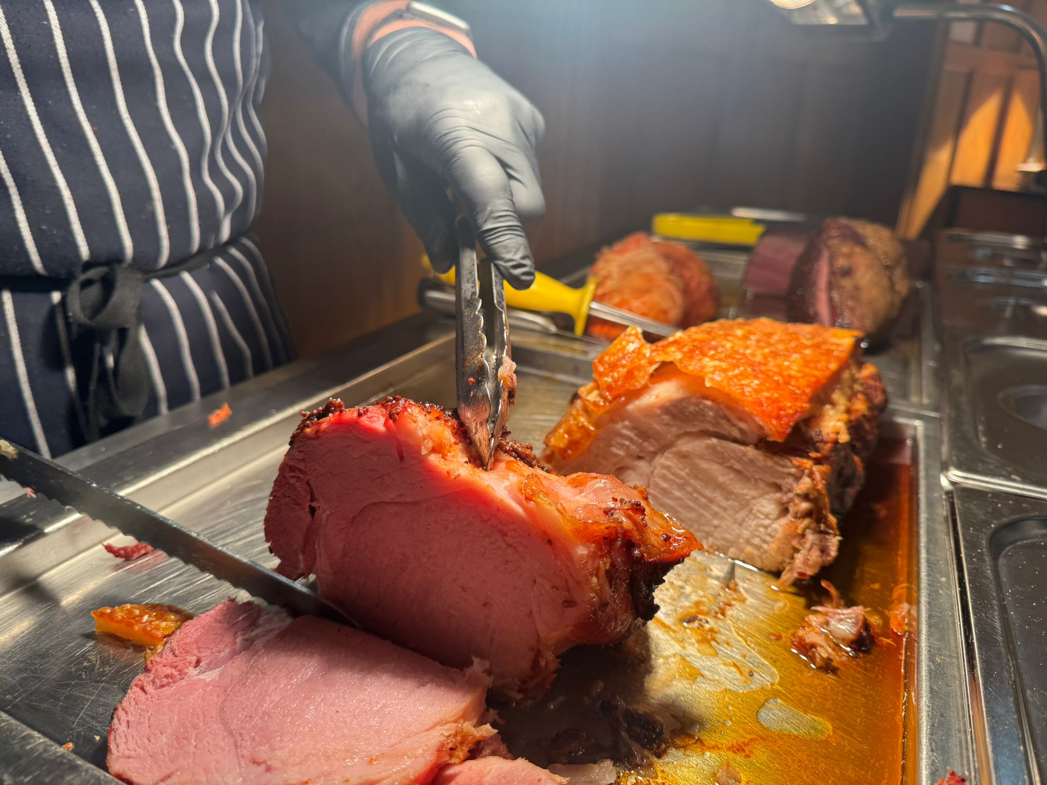 A chef’s hands wearing black gloves, using a carving fork and knife to slice into a succulent, pink roasted joint of meat.